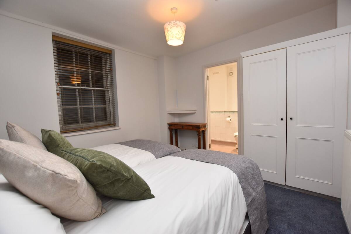 A bedroom with a bed, pillows, a window with blinds, a bedside table, and a wardrobe.