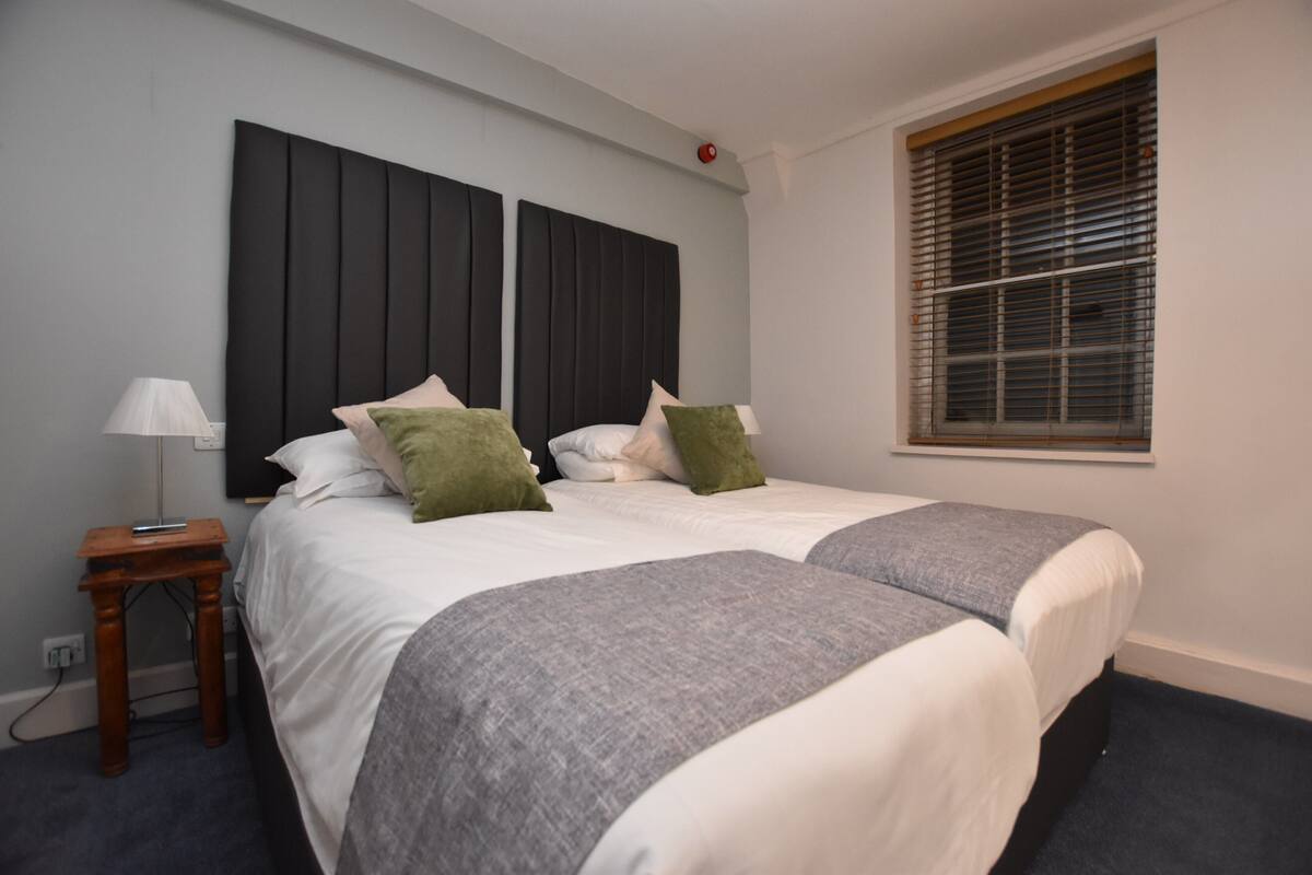 A double bed with two pillows, a bedside table with a lamp, and a window with blinds.