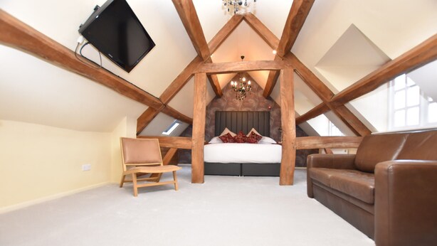 Une chambre avec un plafond en pente, un lit, un canapé en cuir marron, une chaise en bois et un téléviseur à écran plat fixé au mur.