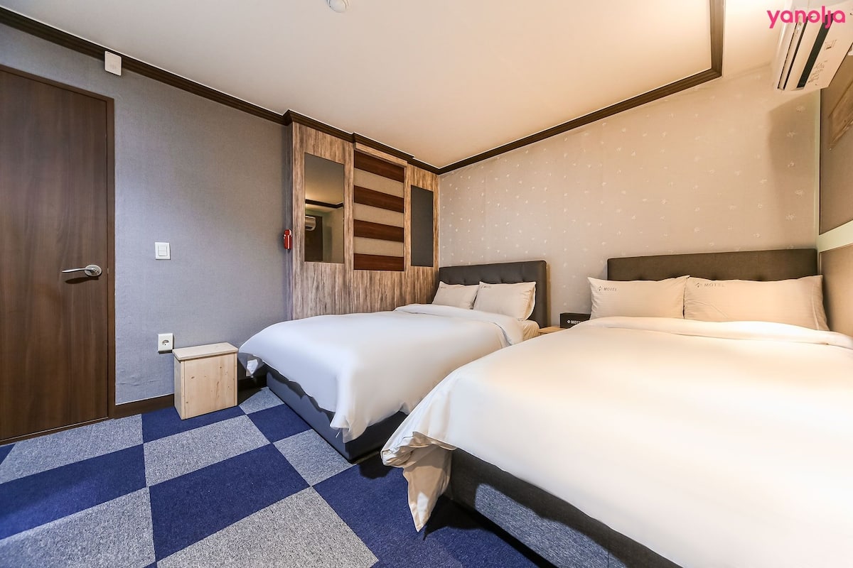 A hotel room with two beds, a wooden wardrobe, a small bedside table, and a patterned carpet.
