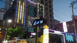 Un bâtiment avec une enseigne au néon indiquant un motel, un taxi garé devant et un panneau avec un symbole représentant un pouce levé.
