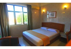 A hotel room with a bed, a bedside table, a chair, a window with curtains, and a wall-mounted light fixture.