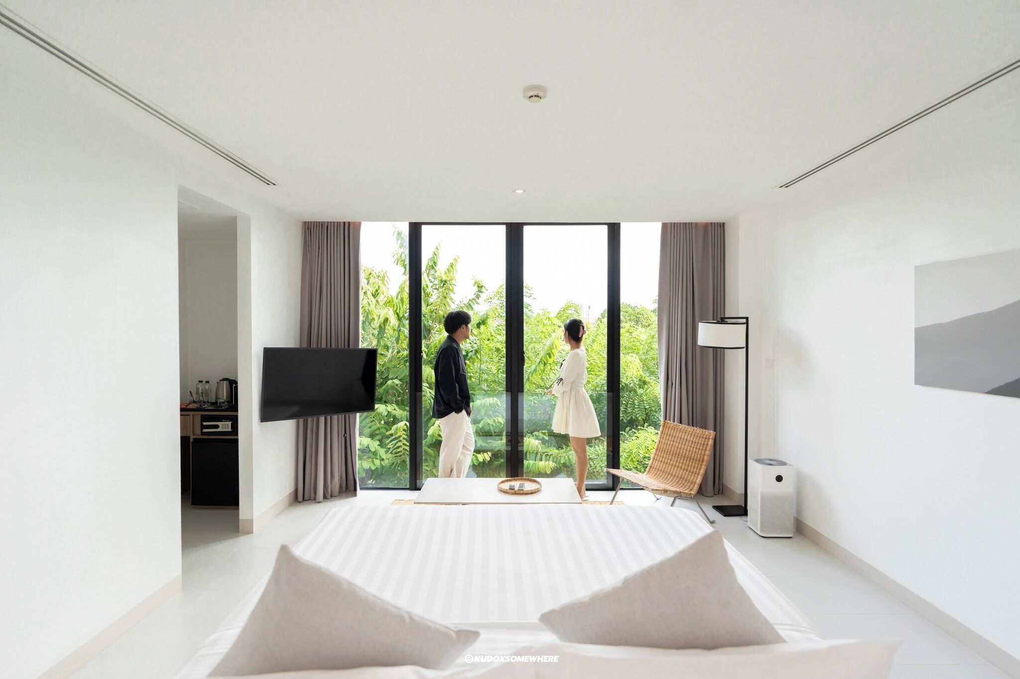A modern bedroom with a large bed, a flat-screen TV, and a view of greenery through large windows.
