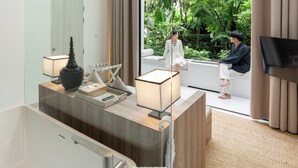 A modern hotel room with a large window, a wooden vanity, a flat-screen TV, and a seating area with a view of lush greenery. - K Maison Lanna Boutique Hotel (Chiang Mai)