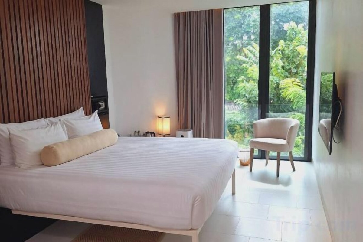 Un dormitorio con una cama grande, una mesita de noche, una silla y vistas a la vegetación por la ventana.