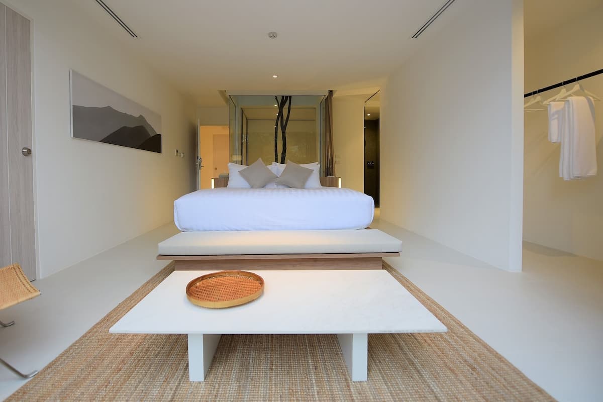 A modern hotel room with a minimalist design, featuring a large bed, a small table, and a view of the hallway.