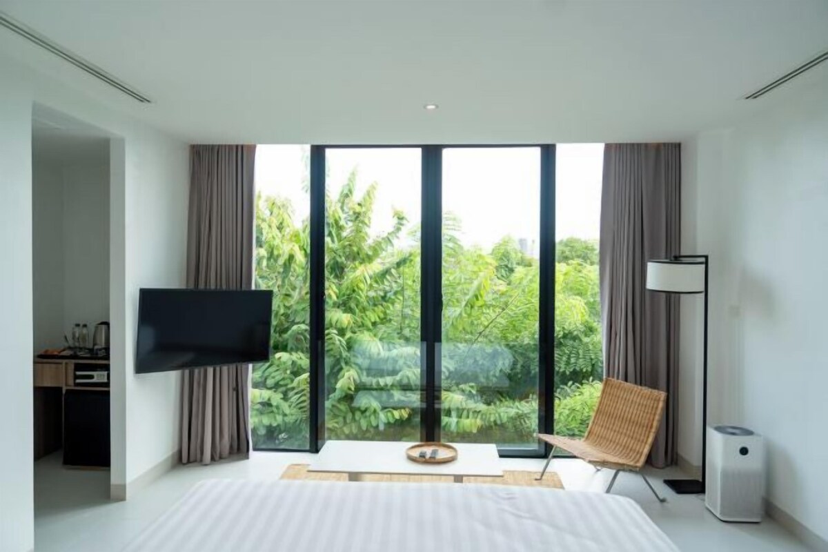 A modern hotel room with a large bed, a flat-screen TV, a desk with a computer, and a view of lush greenery through floor-to-ceiling windows.