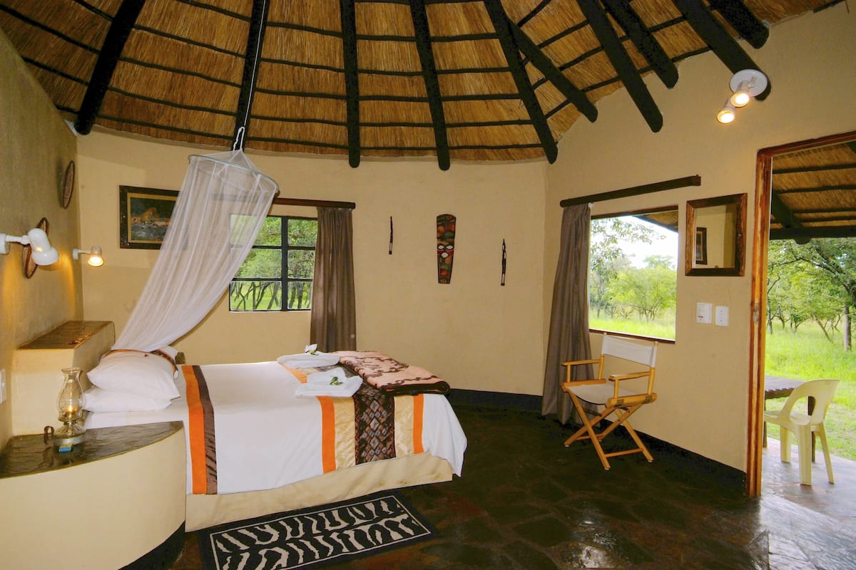 A room with a bed, a chair, a bedside table, a lamp, a mosquito net, and a window with curtains.