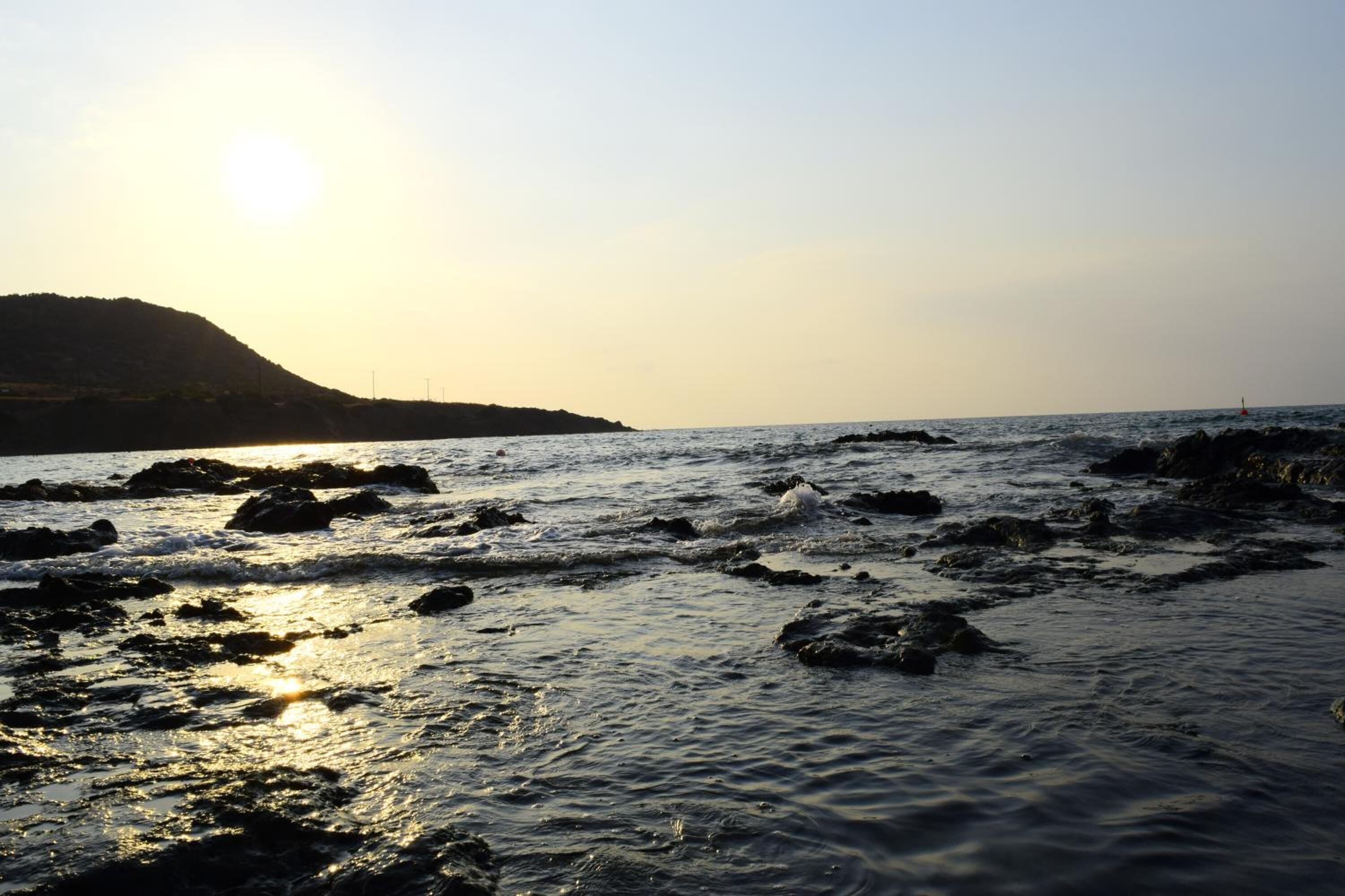 A beach with rocks and calm water, a clear sky, and a distant hill.