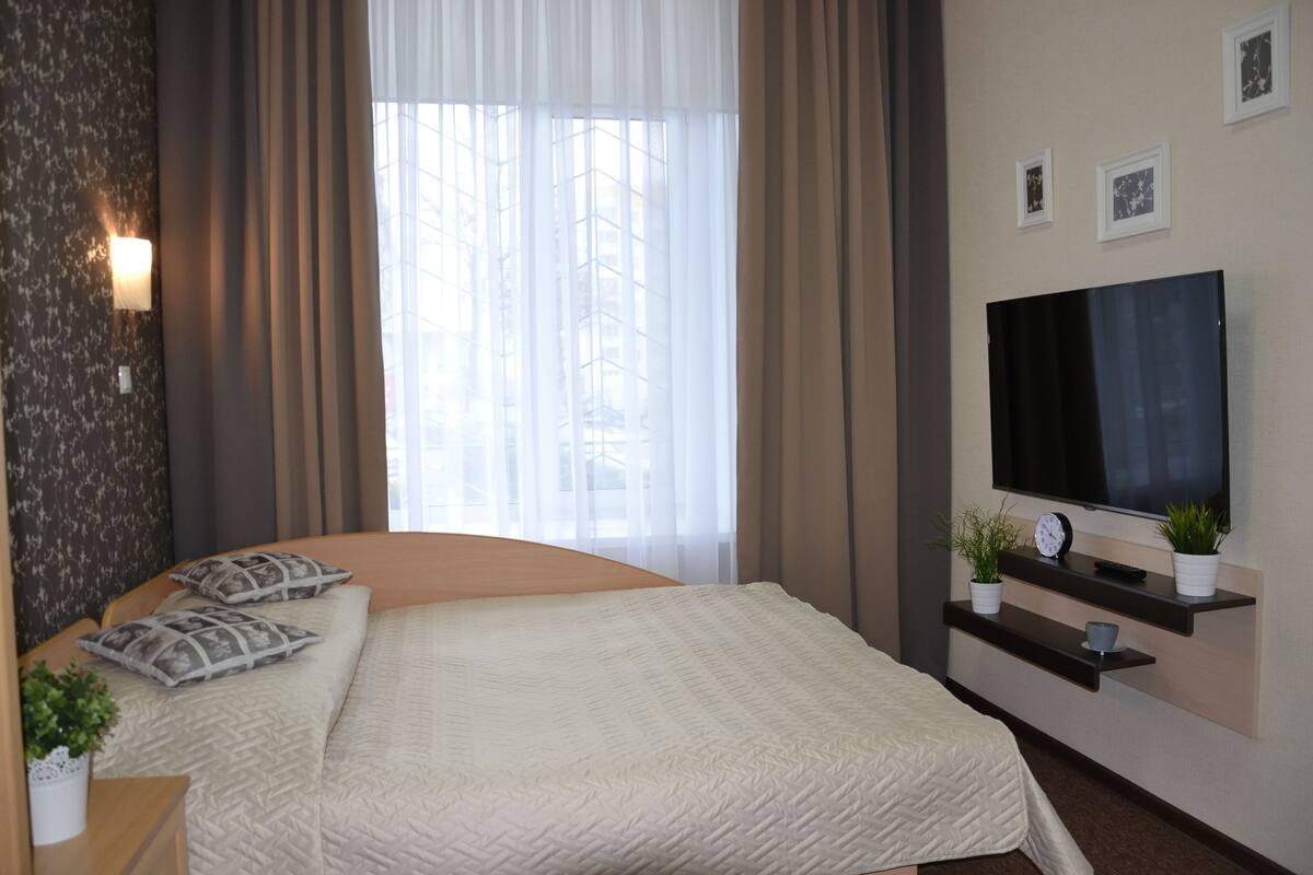 A modern hotel room with a large bed, a flat-screen TV, a bedside table with a lamp, and a window with sheer curtains.