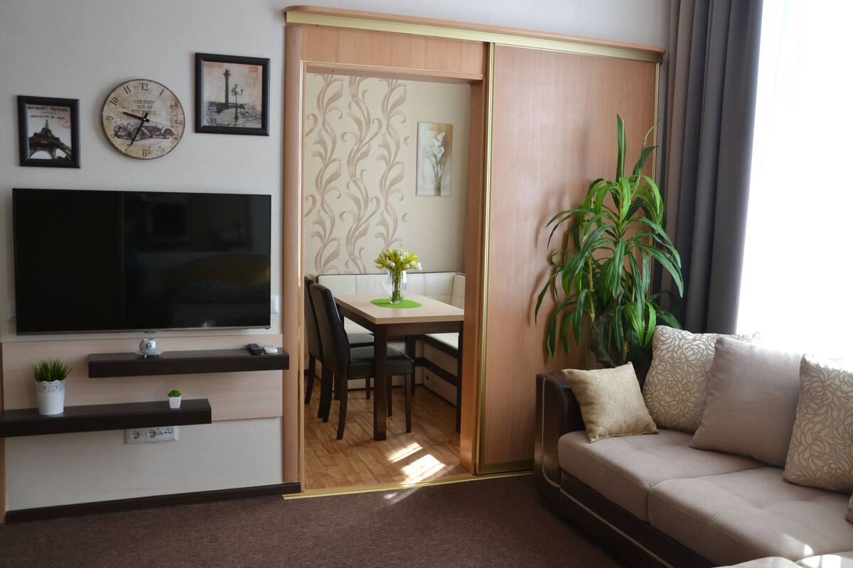 A living room with a TV, a dining table, chairs, a sofa, a plant, and a sliding door.