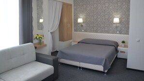 Une chambre d’hôtel moderne comprenant un lit, un canapé, une table de chevet avec une lampe, un miroir et un mur recouvert d’un papier peint à motifs.