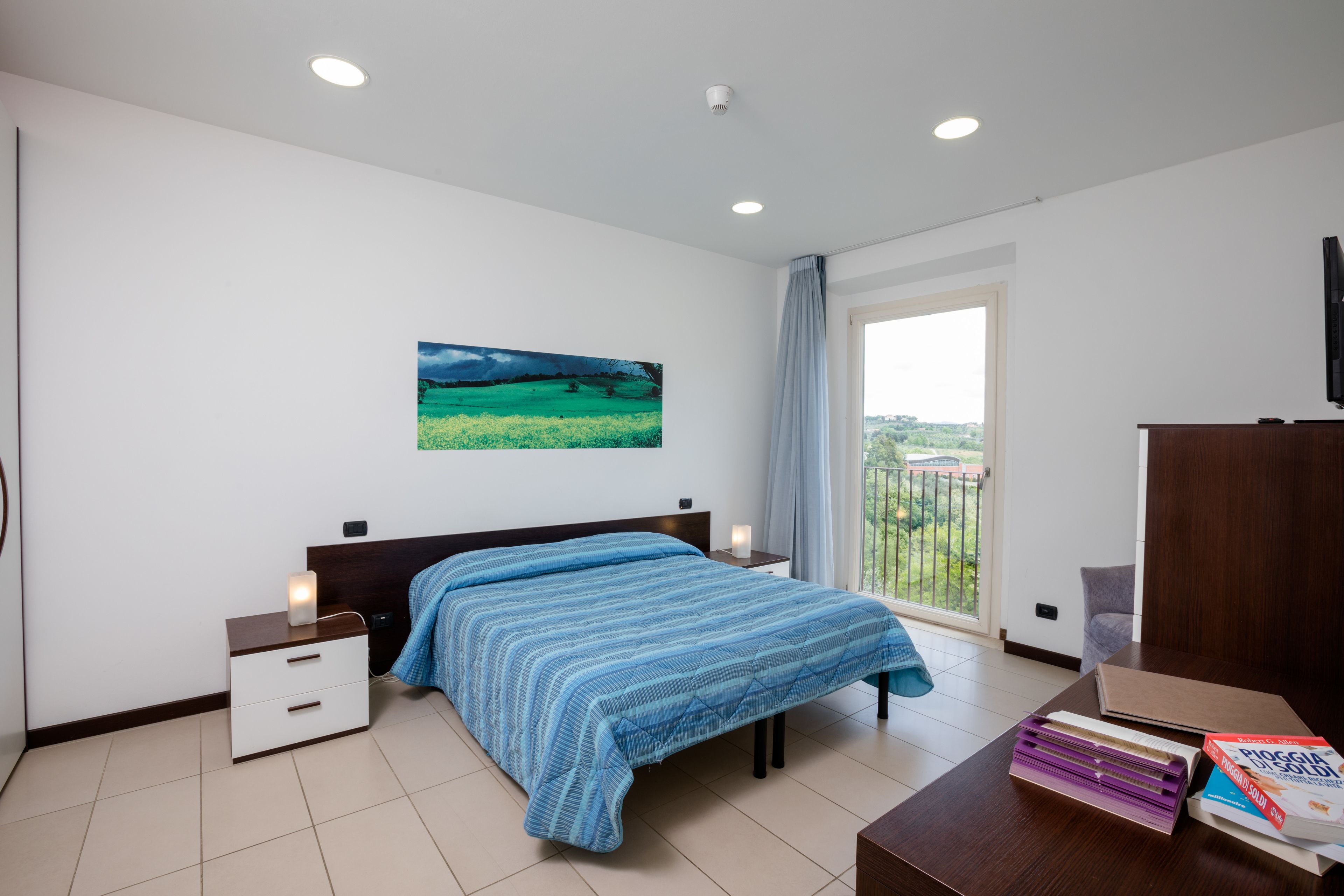 A bedroom with a bed, bedside tables, a painting, a window with curtains, and a view of greenery.