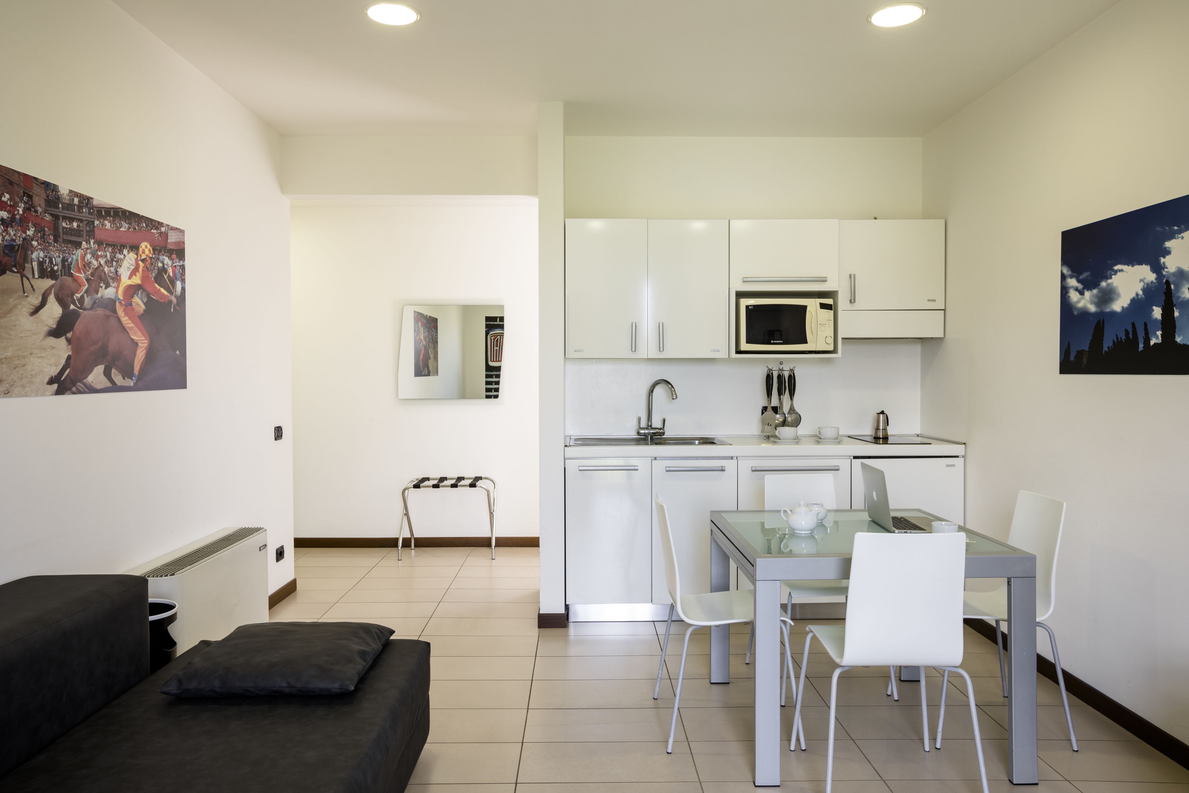 A compact kitchen with white cabinets, a microwave, and a sink. There is a dining table with chairs and a black couch with a pillow.