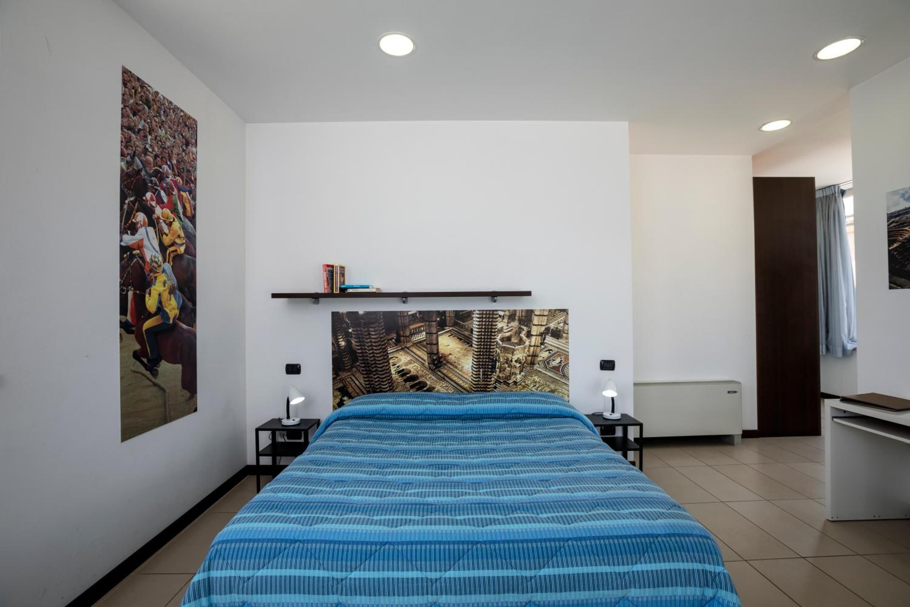 A bedroom with a bed, a wall-mounted shelf with books, a large painting, and a desk with a lamp.