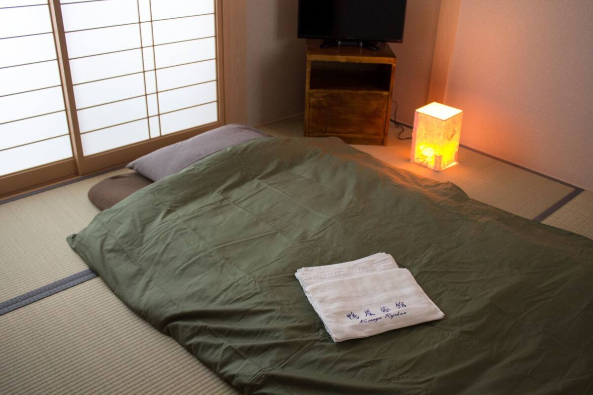 Una habitación con suelo de tatami, una cama con edredón verde, una toalla blanca con texto, un televisor sobre un soporte de madera y una lámpara de pie encendida.