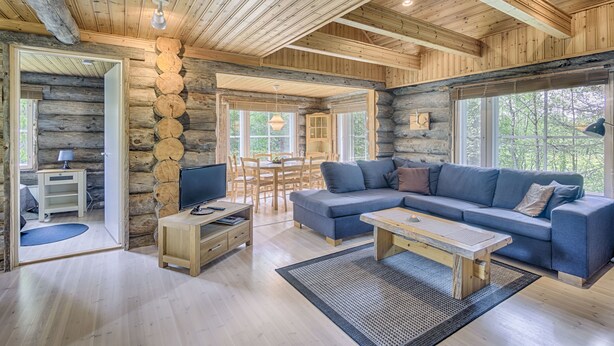 Ein Interieur einer Blockhütte mit Holzbalkendecke, einer Couch, einem Couchtisch, einem Fernseher und einem Essbereich.