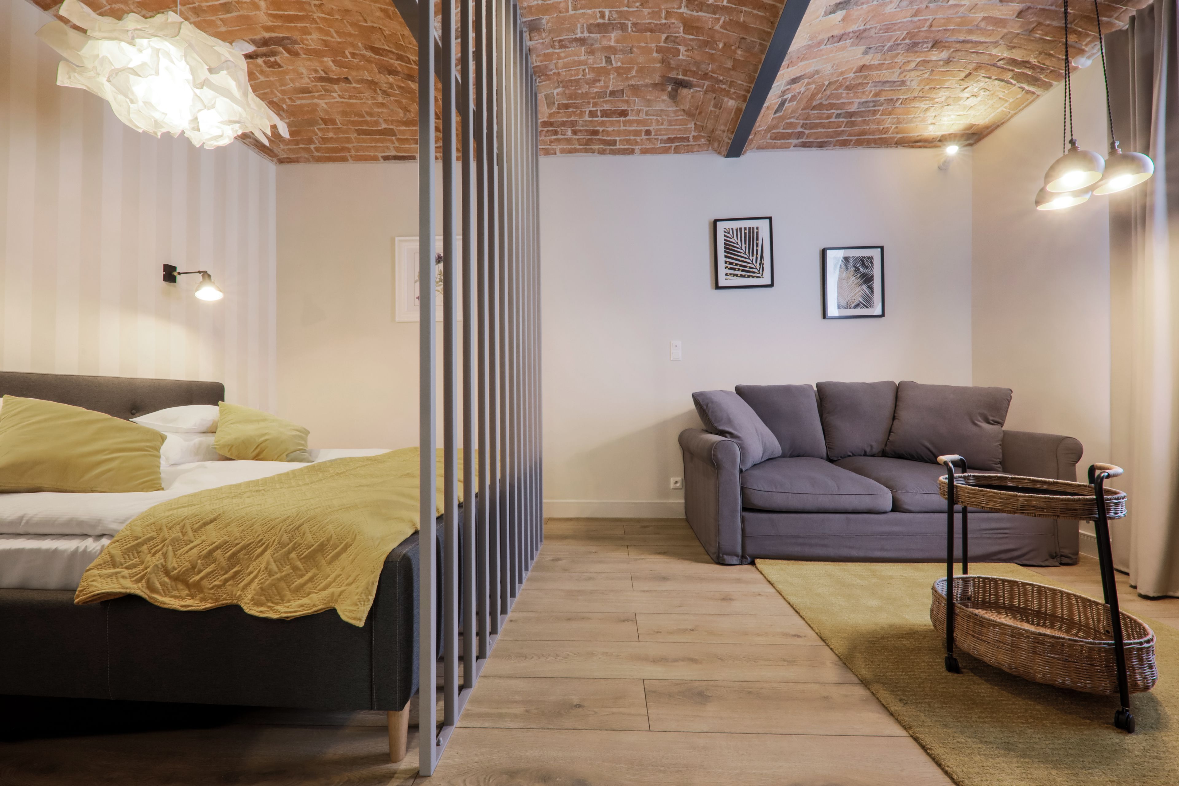 A room with a bed, a grey sofa, a wicker side table, and a brick ceiling.