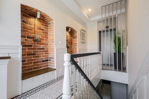 Uma escadaria com balaústres brancos e corrimão preto, que leva a um patamar com parede de tijolos e um lustre suspenso.