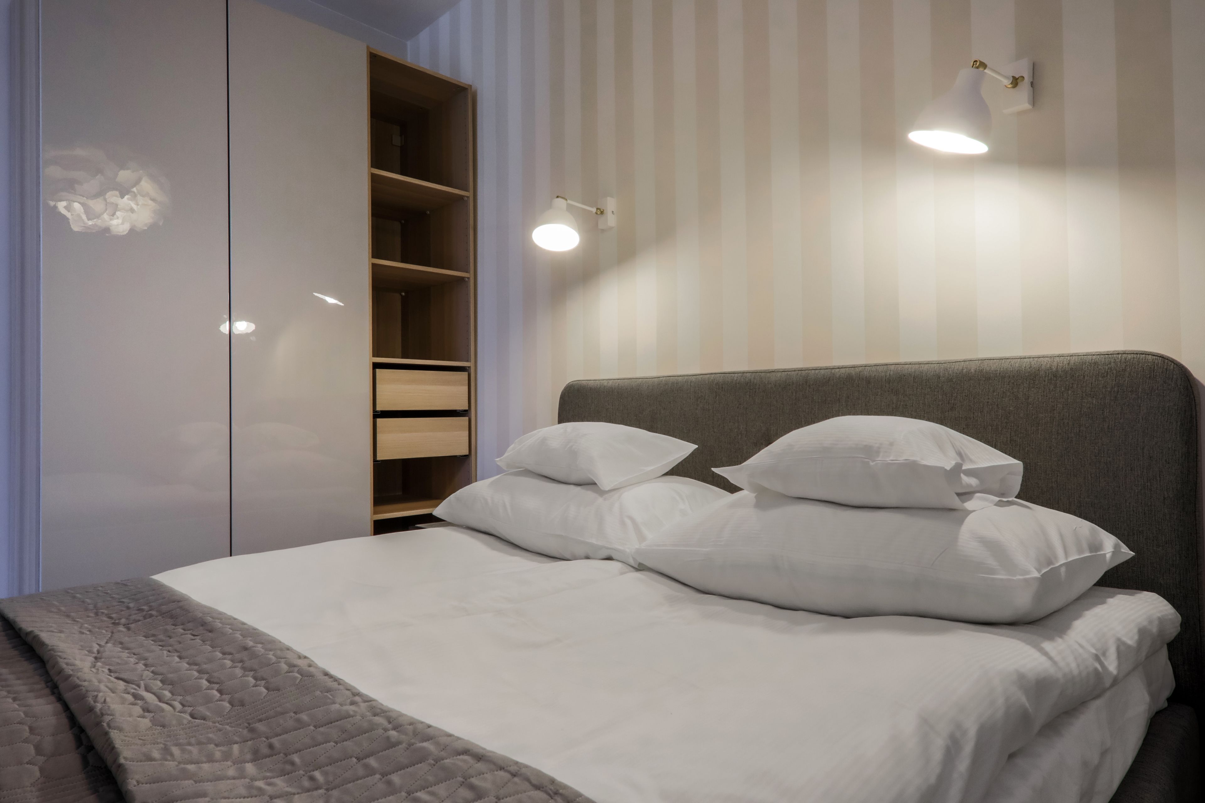 Una cama bien hecha con sábanas y almohadas blancas, un cabecero gris y un armario con estantes abiertos.