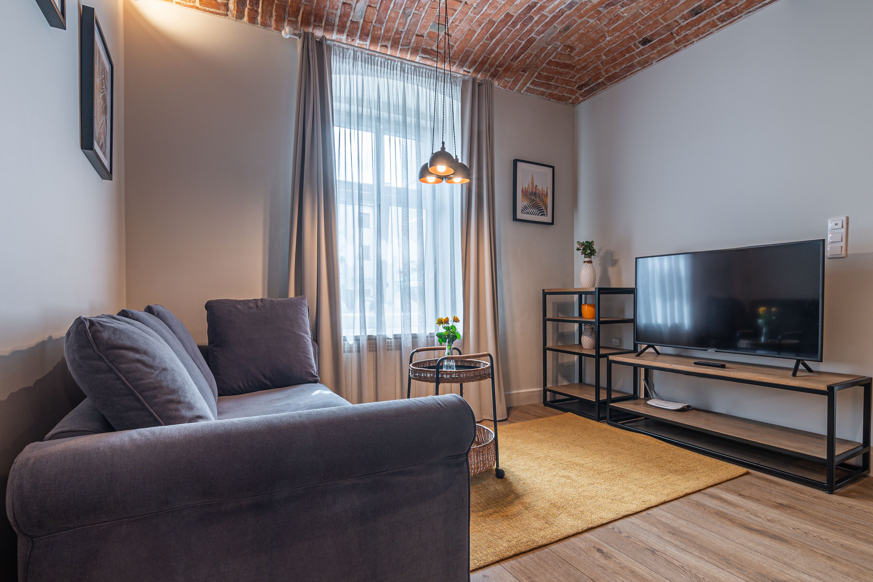 Een moderne woonkamer met een grijze bank, een televisie op een houten dressoir en een metselwerk plafond.