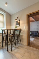 Uma sala de jantar com mesa e cadeiras de madeira, um quadro na parede, e vista para uma sala de estar com parede de tijolos e um sofá.