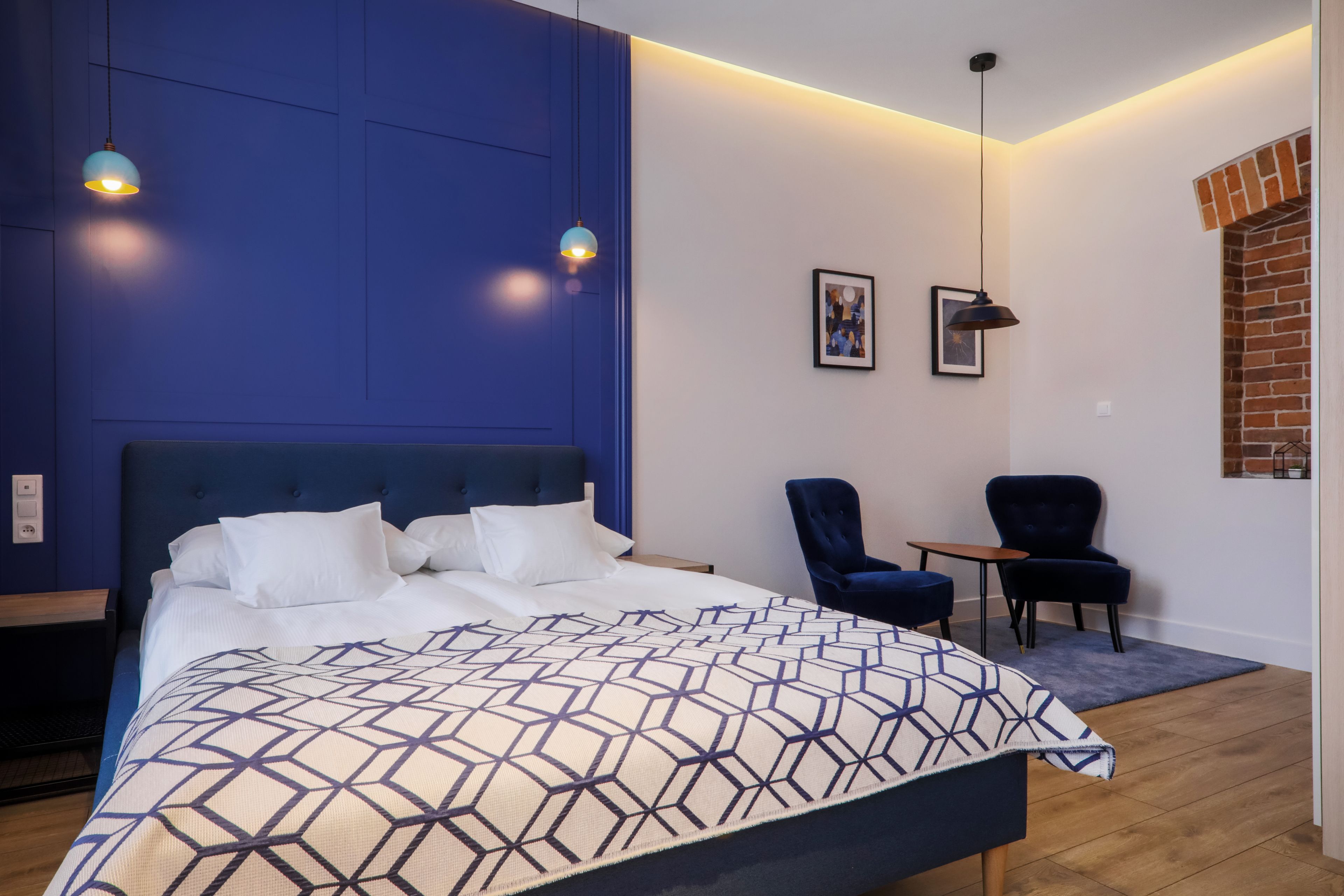 A modern bedroom with a large bed, two pendant lights, a small table, two chairs, and a brick fireplace.