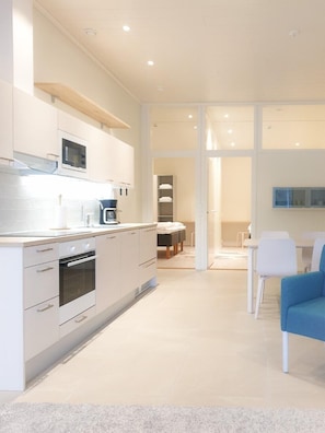A modern kitchen with white cabinetry, a built-in oven, and a microwave. There is a dining area with a white table and chairs, and a blue chair.
