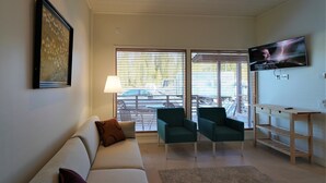 Una sala de estar con un sofá, dos sillones, un televisor montado en la pared y una ventana con persianas.