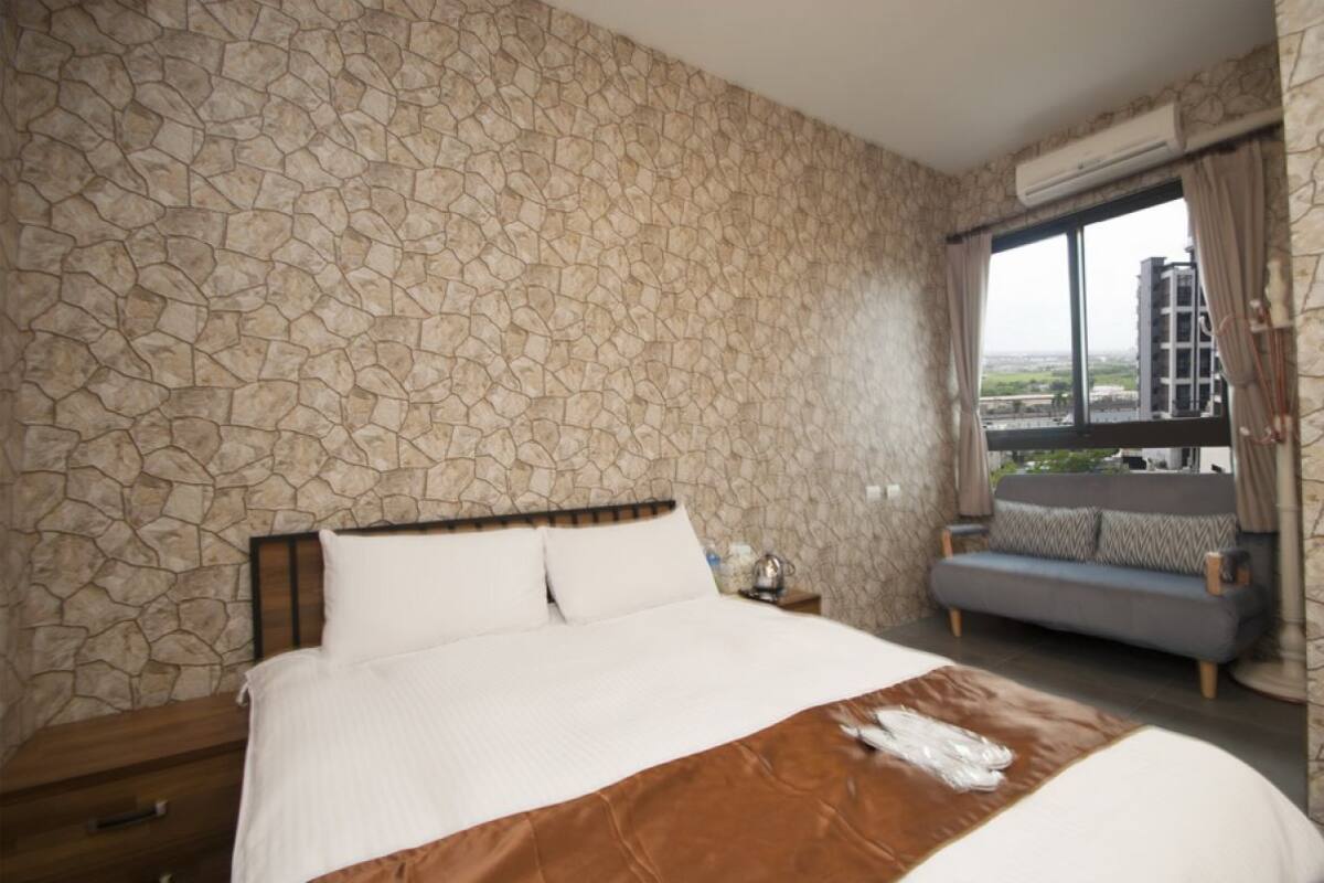 A bedroom with a stone-patterned wall, a bed with white and brown bedding, a nightstand, a sofa, and a window with a view of buildings.