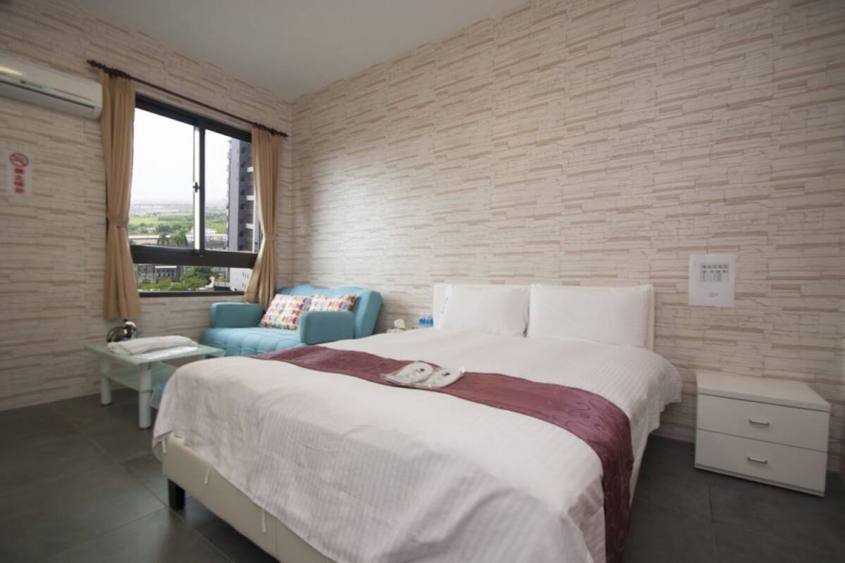 A hotel room with a large bed, a small table, a chair, and a window with curtains.