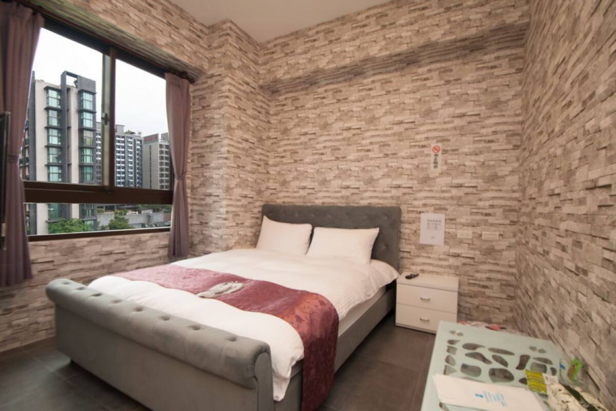 A bedroom with a bed, a nightstand, a window, and a brick wall.