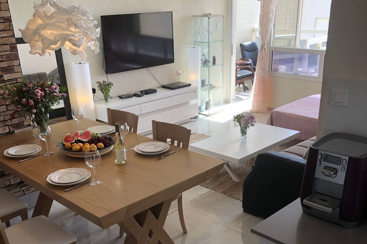 Uma área de jantar moderna com mesa de madeira posta para quatro pessoas, uma TV de tela plana na parede e uma cozinha com máquina de café.