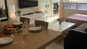 Un salon moderne avec une table à manger en bois dressée pour le repas, un téléviseur à écran plat fixé au mur et une cloison en verre séparant l’espace repas du salon.