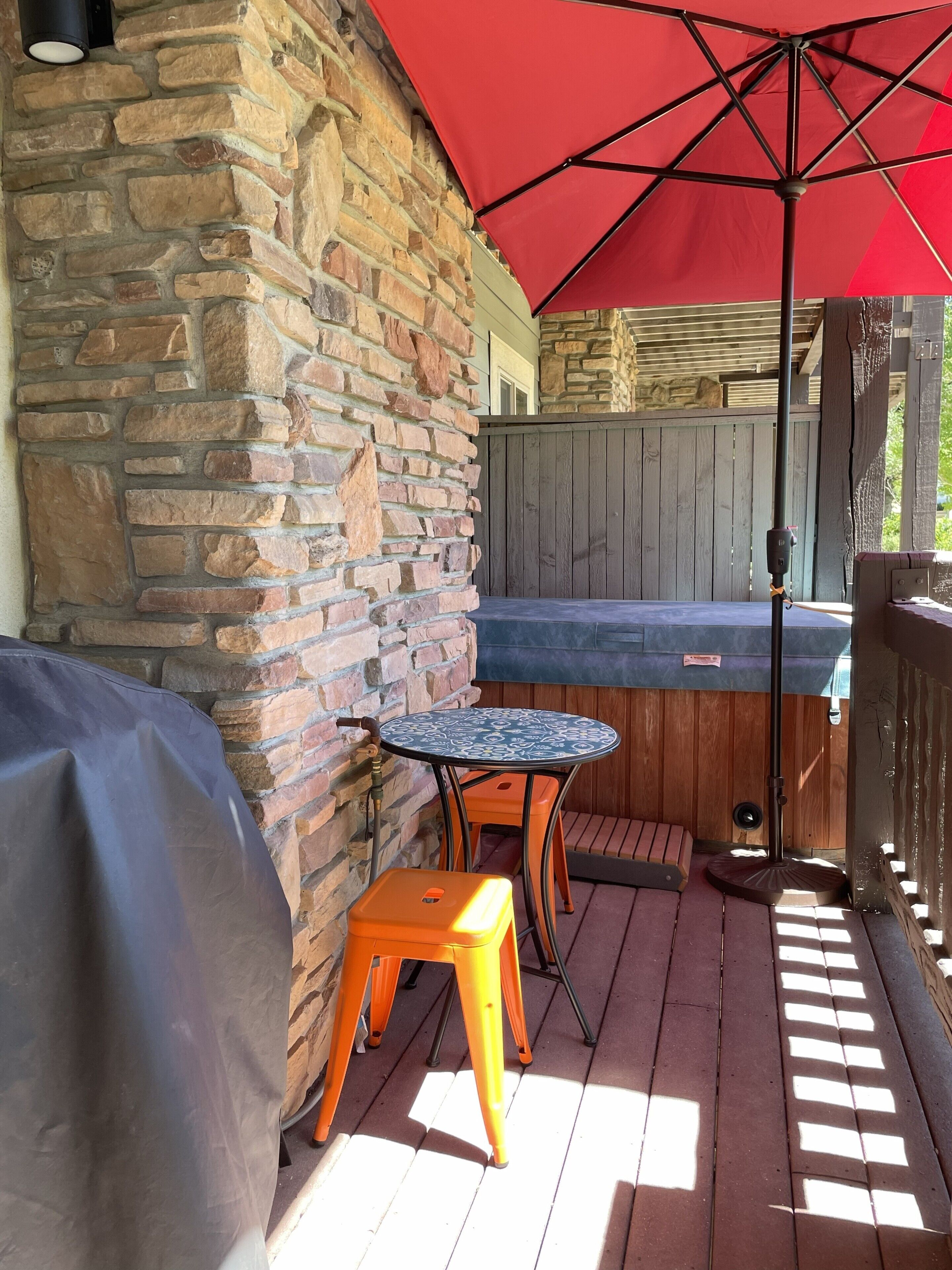 Un'area patio con un muro in pietra, una zona relax coperta con tavolo e sedie, e un ombrellone rosso che offre ombra.