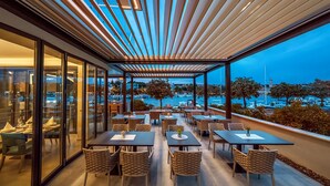 Un área de comedor al aire libre con mesas y sillas, con vista a un puerto deportivo y barcos.