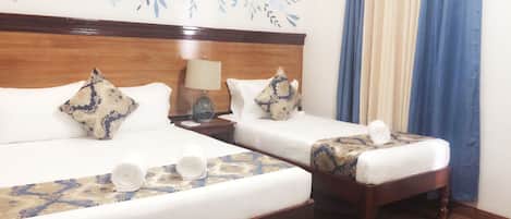 A hotel room with two beds, a wooden headboard, a nightstand with a lamp, and a window with blue curtains.