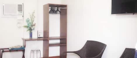 Ein Zimmer mit Holzboden, Schreibtisch mit Stuhl, Kleiderschrank, zwei Korbstühle mit blauen Kissen und einem an der Wand befestigten Flachbildfernseher.