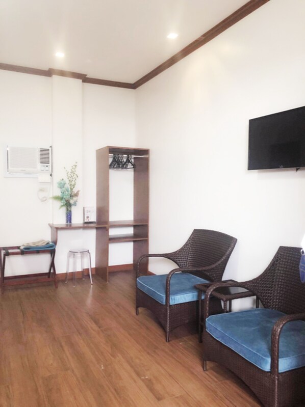 Una habitación con piso de madera, un escritorio con silla, un armario, dos sillas de mimbre con cojines azules y un televisor de pantalla plana montado en la pared.