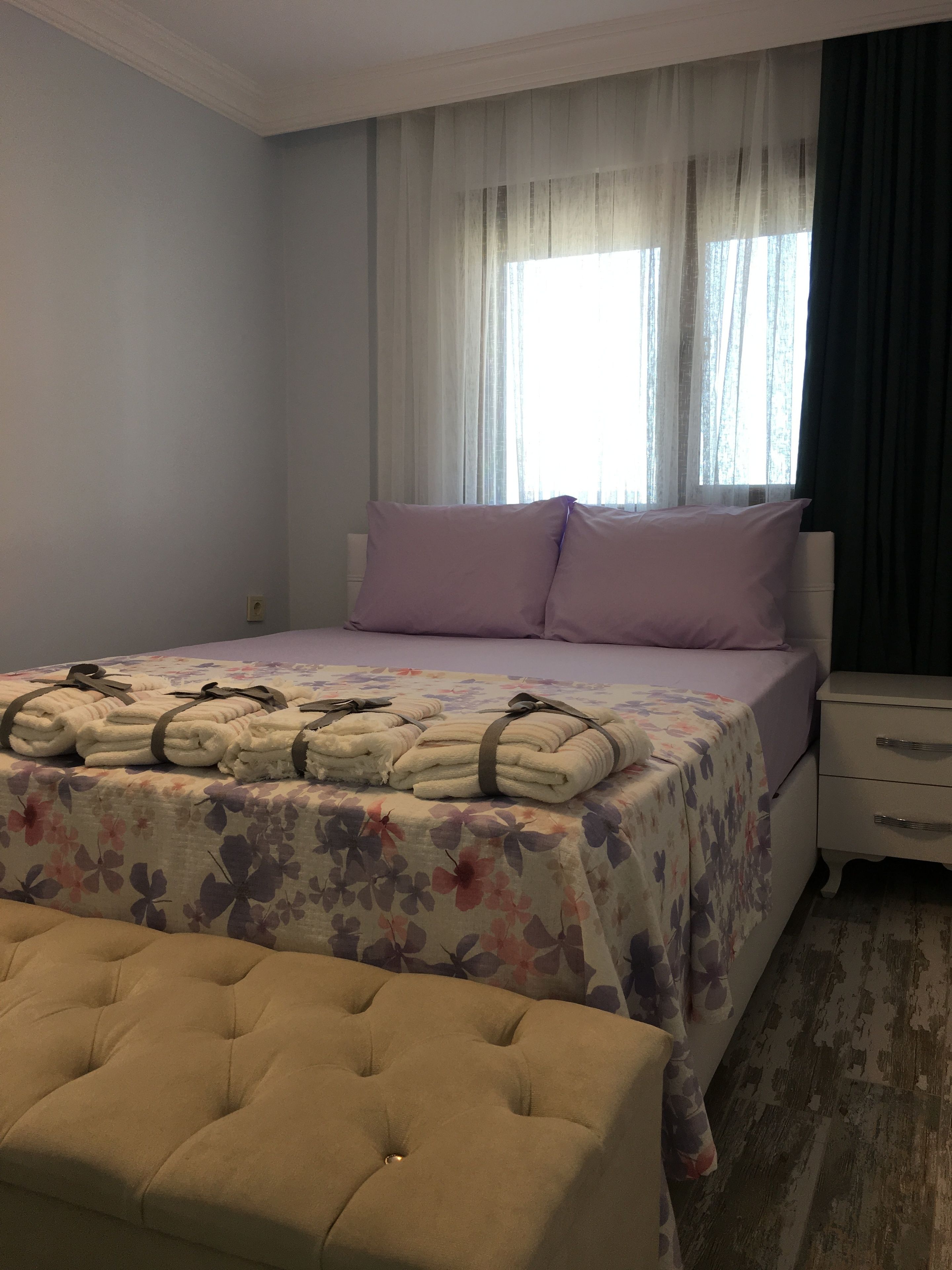 A neatly made bed with a floral bedspread, a bedside table with drawers, and a tufted ottoman.