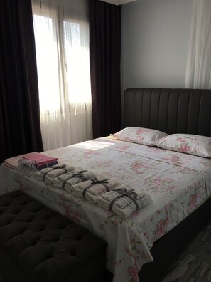 A neatly made bed with floral bedding, a headboard, and a bedside table with a pink blanket and a folded towel. - Seraysen Apart (Seferihisar)