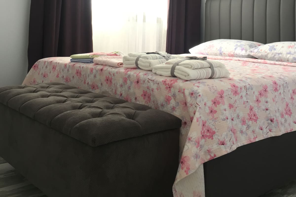 A neatly made bed with a floral bedspread, a grey upholstered bench, and a window with dark curtains.
