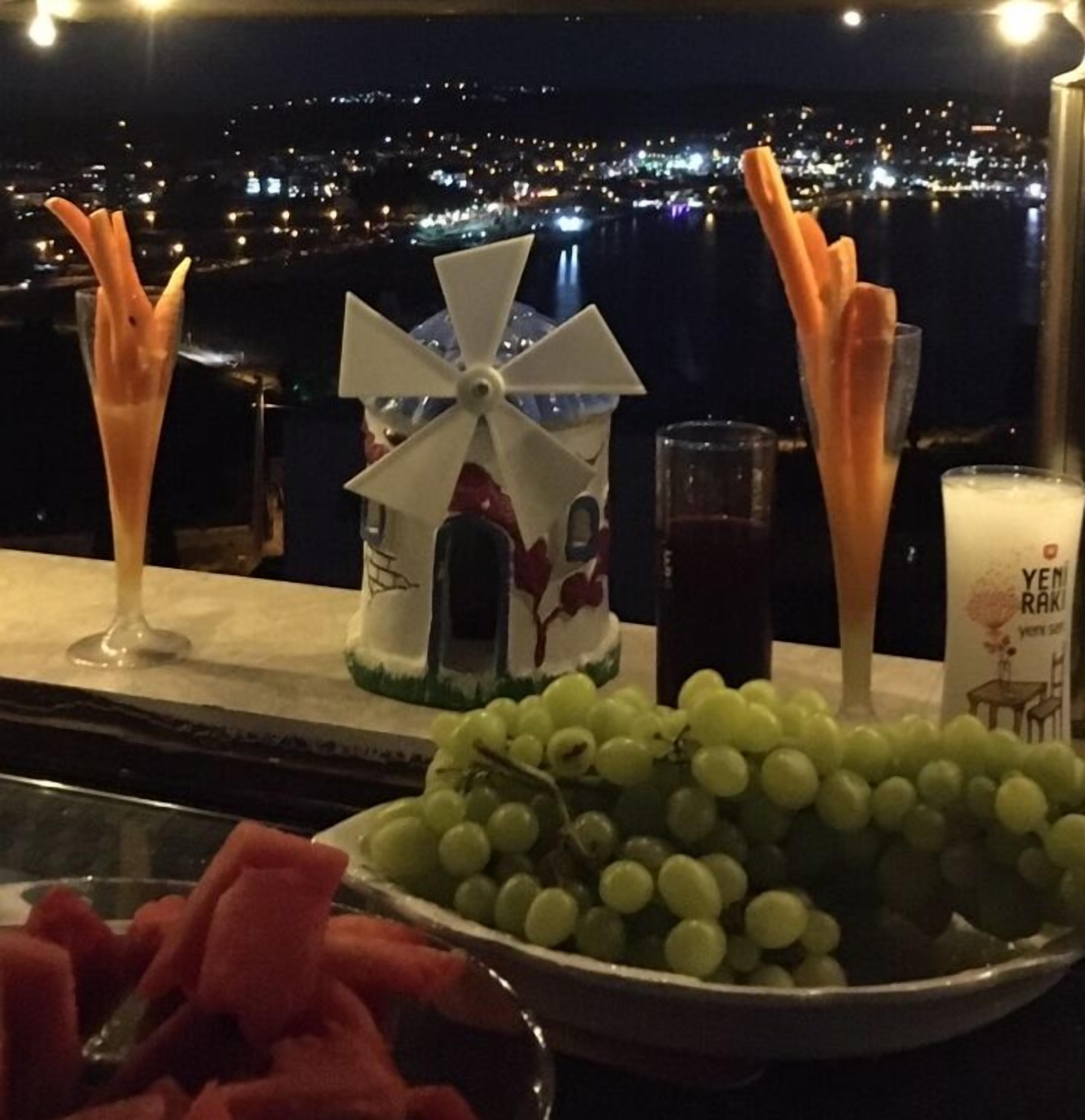 Une table avec un bol de raisins, un verre à vin et une bougie portant l’inscription « YEMRAKI », le tout placé devant un paysage urbain nocturne.