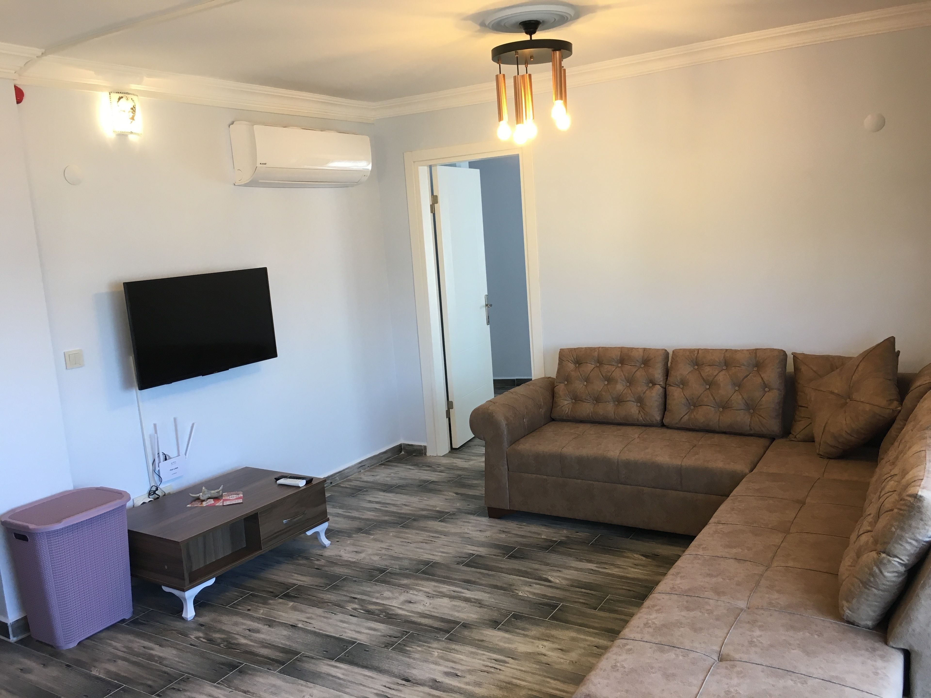 Una sala de estar con un sofá marrón, un televisor de pantalla plana montado en la pared, una pequeña mesita de madera y un cubo de basura.