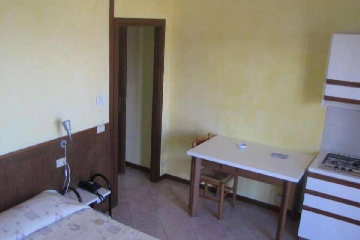 Quarto pequeno e funcional com cama, escrivaninha e cadeira. Possui janela com vista para um edifício.