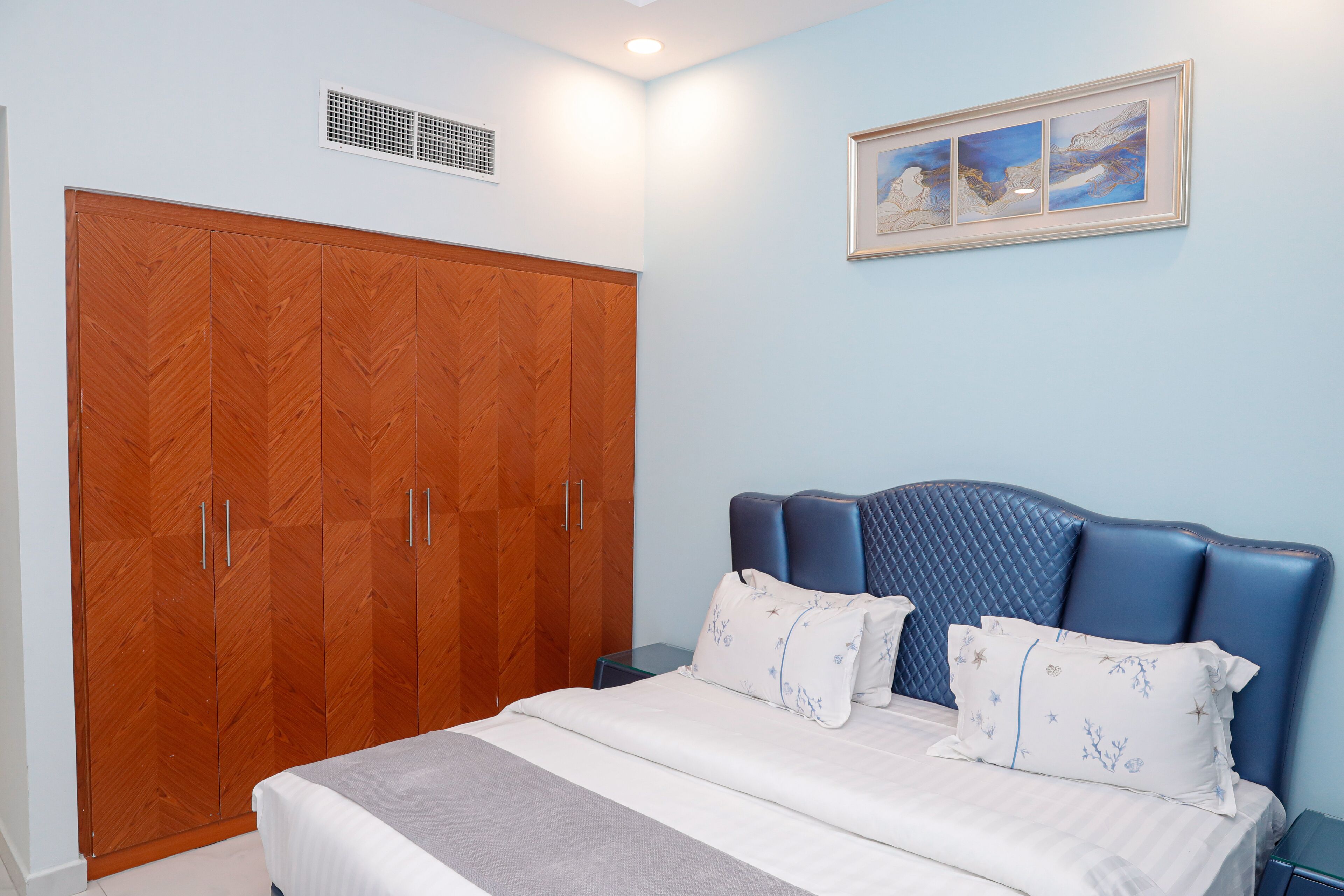 A bedroom with a large wooden wardrobe, a bed with blue headboard, and a framed picture on the wall.