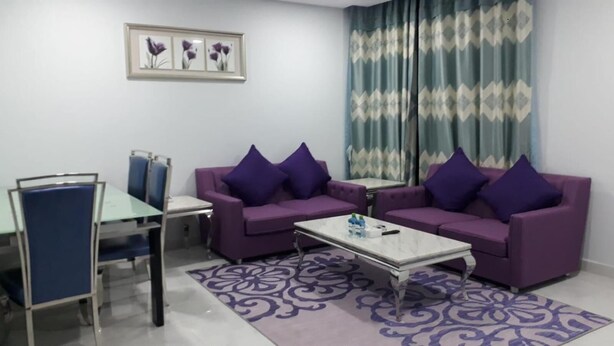 Um quarto com dois sofás roxos, uma mesinha, uma cadeira e cortinas estampadas.