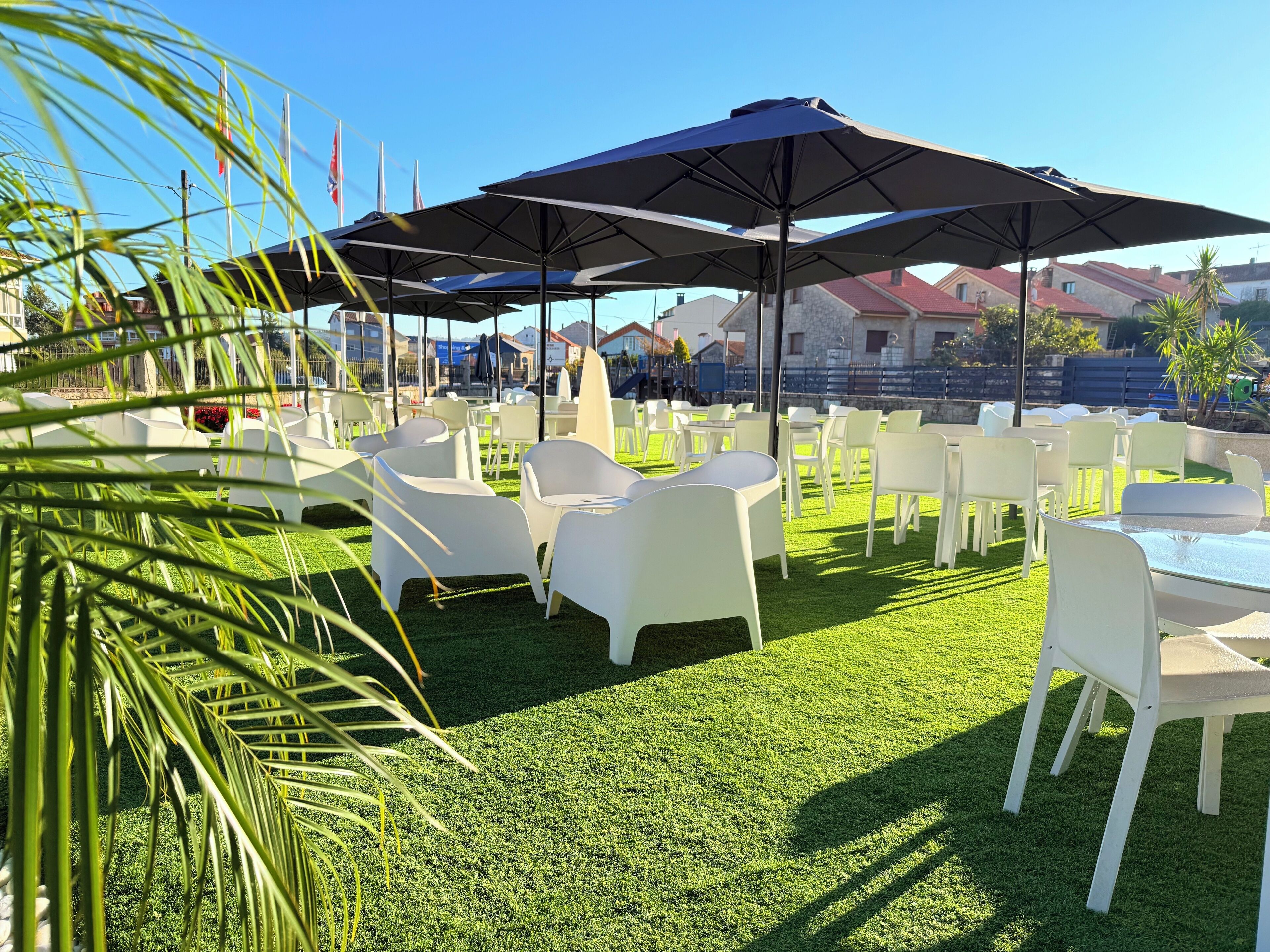 An outdoor seating area with white plastic chairs and tables, shaded by large umbrellas, on a well-maintained lawn.
