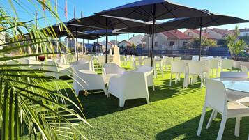 An outdoor seating area with white plastic chairs and tables, shaded by large umbrellas, on a well-maintained lawn.