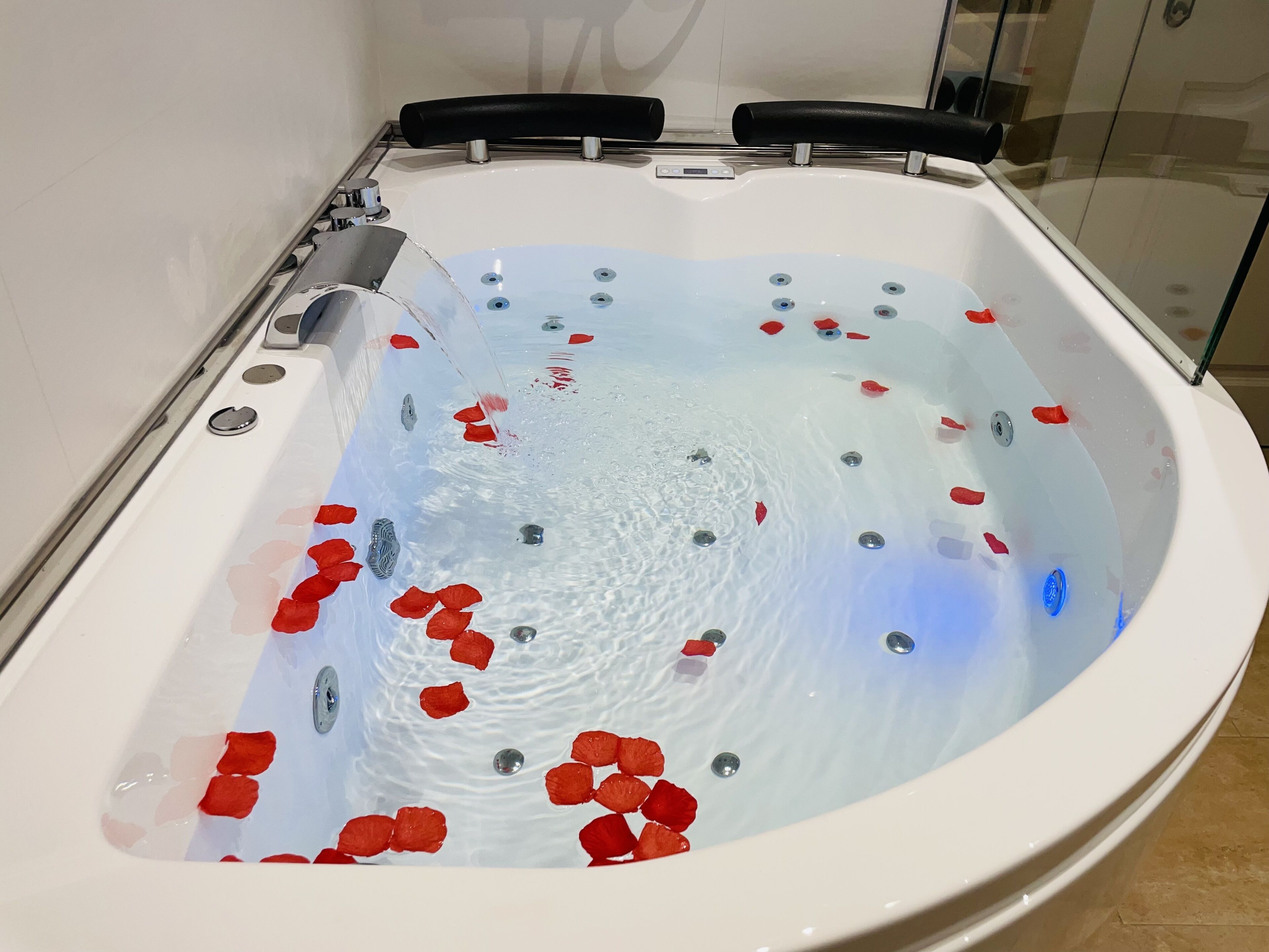 Een modern bubbelbad met rozenblaadjes in het water, een bedieningspaneel en een glazen ombouw.